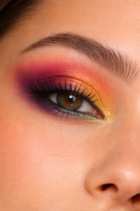 Close-up de olho com maquiagem esfumada em sombras vibrantes, em tons quentes de laranja, rosa e roxo, com acabamento suave e natural.