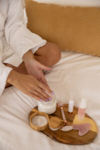 Mãos delicadas tocam um pote de creme sobre uma bandeja de madeira com produtos de skincare, como sérum, hidratante e rolinho facial de quartzo rosa, representando cuidados diários com a pele em um ambiente suave e acolhedor.