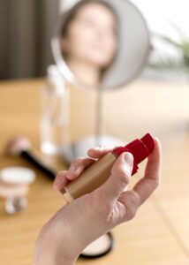 Mão segurando um frasco de base líquida com tampa vermelha, pronta para aplicação, com espelho e itens de maquiagem desfocados ao fundo, ilustrando como escolher a base perfeita.