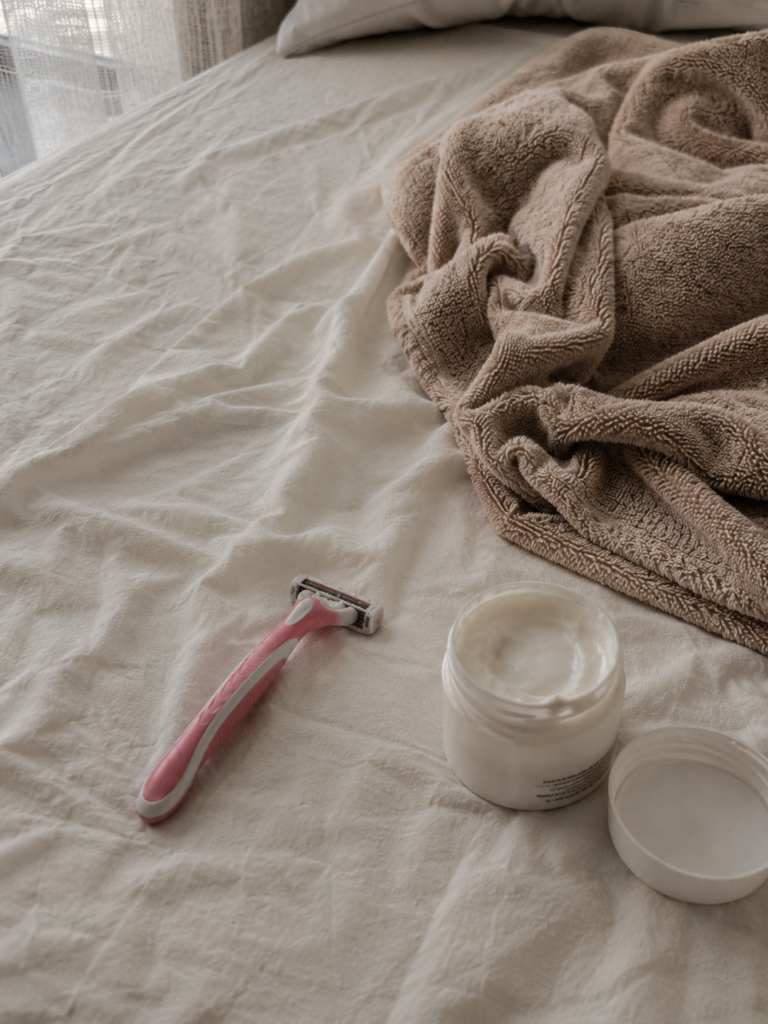 Lâmina de depilação e creme hidratante sobre uma cama com lençol claro e toalha bege, iluminados por luz natural.