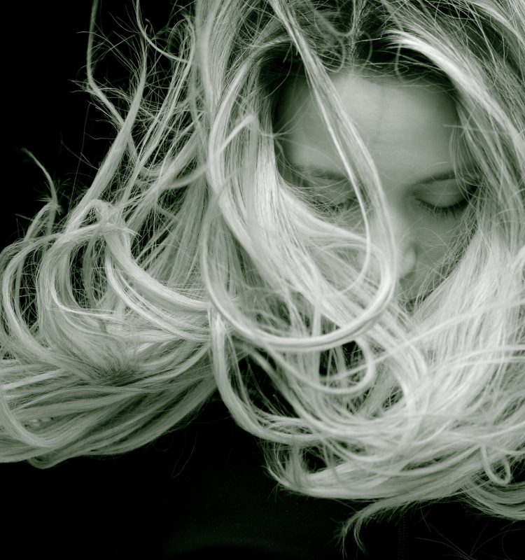 Mulher de cabeça baixa, cabelos longos com raiz escura e luzes marcantes, em fundo preto, fios em movimento que transmitem leveza, harmonia e presença delicada.