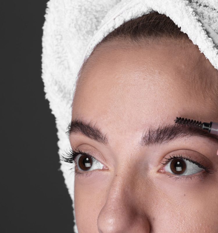 Pessoa escovando as sobrancelhas diante do espelho, com os cabelos envolvidos em uma toalha, em um gesto calmo de cuidado cotidiano com o olhar.