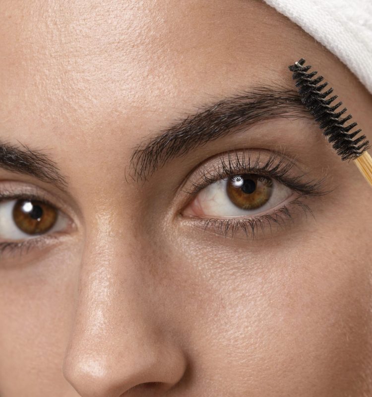 Close de uma mulher penteando as sobrancelhas com escovinha, com foco nos olhos castanhos e na pele com textura natural.