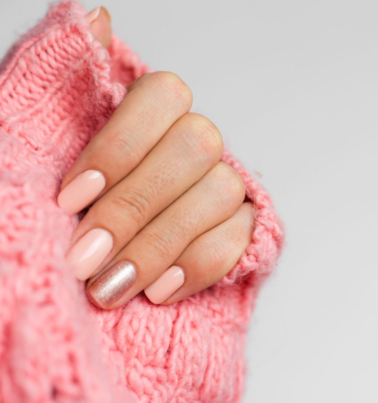 Mão com unhas rosadas em tom suave, acabamento delicado e uma unha com leve brilho acetinado, envolvida por tricô rosa claro, transmitindo cuidado, aconchego e beleza sutil.